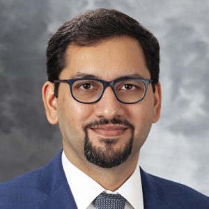 A grey photography background with a formal headshot photo of Muhammed Murtaza, MBBS, PhD, wearing a dark blue suit and tie.