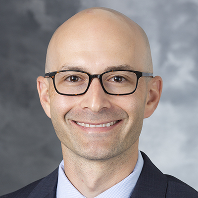 A headshot of David Harris, MD, who is wearing a dark grey suit jacket, a blue dress shirt, and black and brown frame glasses.