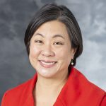 A grey photography background with a formal photo of Sandra Lin, MD, smiling with black hair and a red jacket.