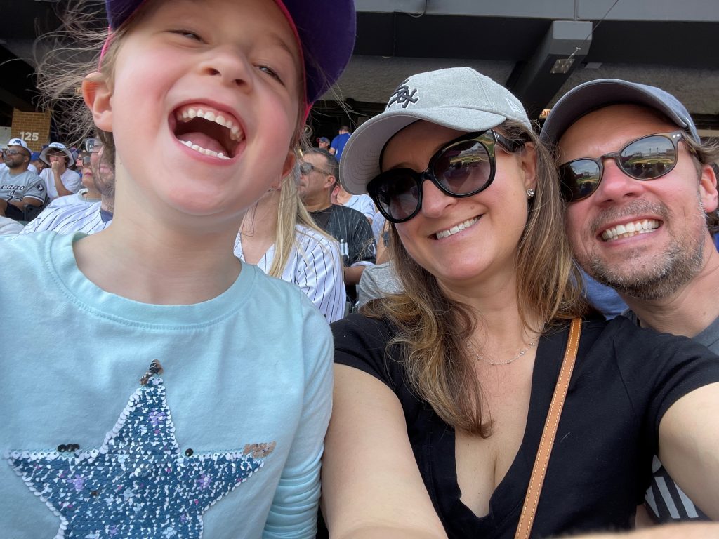 Dr. Summer Hanson, wearing a White Sox hat and sunglasses, smiles with her daughter, Rori, in a blue shirt with a star on it and her husband, Dr. Nick Maassen, who is wearing sunglasses and a hat, while sitting at a White Sox game,.