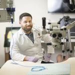 Photo of Dr. Aaaron Dingle in a white lab coat sitting at a lab bench surrounded by microsurgery equipment