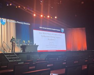 Dr. Sydney Tan stands at a podium at the front of an auditorium. Two people sit at a table next two her. A projector screen has her presentation title slide that says "20th Annual Academic Surgical Congress. Are Work Hours Actually Linked to Burnout Among Surgery Residents?" 