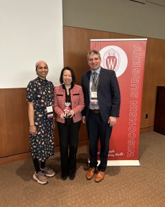 Dr. Mehreen Kisat, Dean Sandra Wong, and Dr. Patrick Varley stand in front of a banner that says "Wisconsin Surgery." Dean Wong is holding a small statue of Bucky Badger.