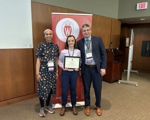 Drs. Mehreen Kisat, Kate Lauer and Patrick Varley stand in front of a banner that says "Wisconsin Surgery." Dr. Lauer holds an award certificate.