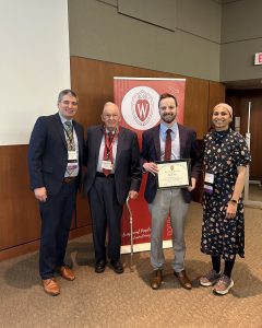 Drs. Patrick Varley and Lou Bernhardt, Daniel Rice, and Dr. Mehreen Kisat stand in front of a banner that says "Wisconsin Surgery." Daniel Rice is holding an award certificate