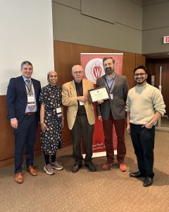 Drs. Patrick Varley, Mehreen Kisat, Layton Rikkers, Matthew Brown, and a member from the Brown Lab stand in front of a banner that says "Wisconsin Surgery." Dr. Rikkers is presenting Dr. Brown with an award