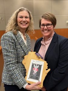 Dr. Rebecca Minter stands next to Dr. Lesly Dosset holding a wooden carving of the state of Wisconsin