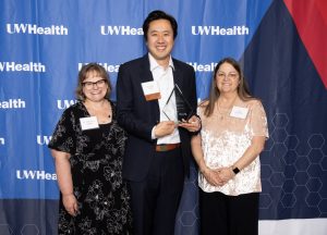 Dr. Rebecca Sippel, Dr. Alex Chiu, and Kar Hamilton stand together in front of a banner that says UW Health. Dr. Chiu is holding an award.