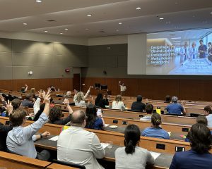 Dr. Jessica Fazendin stands at the front of the auditorium with her hand in the air next to a screen that says "Designing a Clinical Learning Environment That Works Across Generations." Audience members also have their hands in the air.